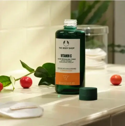 A bottle of The Body Shop Vitamin C Glow Revealing Tonic on a tiled surface with greenery and cotton pads in the background.