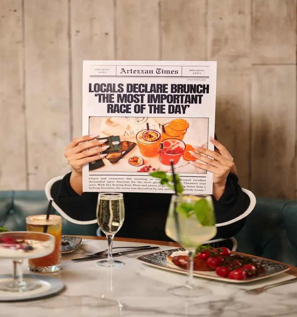A person holds a newspaper titled "Artezian Times" while brunch items and cocktails are displayed on the table in a stylish setting.