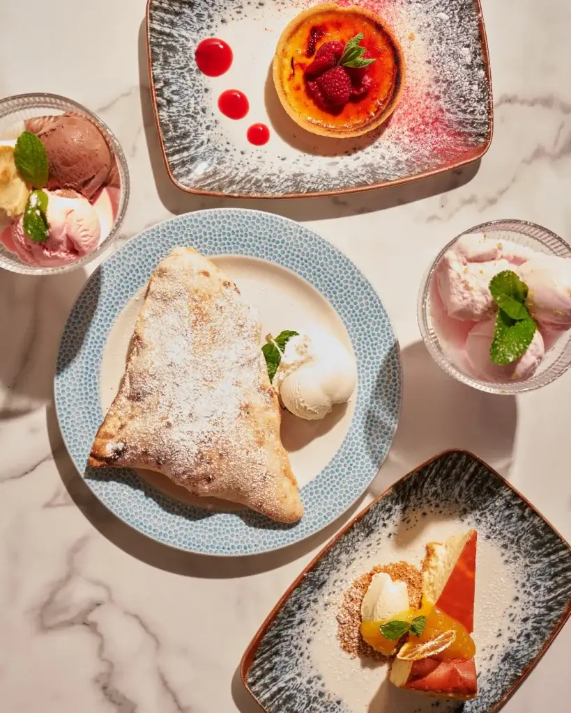 A delicious assortment of desserts: a creamy flan, fruit-topped cheesecake, a powdered pastry with ice cream, and a colorful gelato.