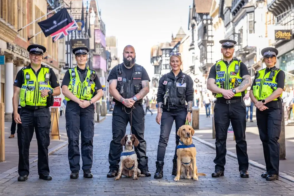 A group of police officers stands on a cobbled street, accompanied by two police dogs, under a clear blue sky.