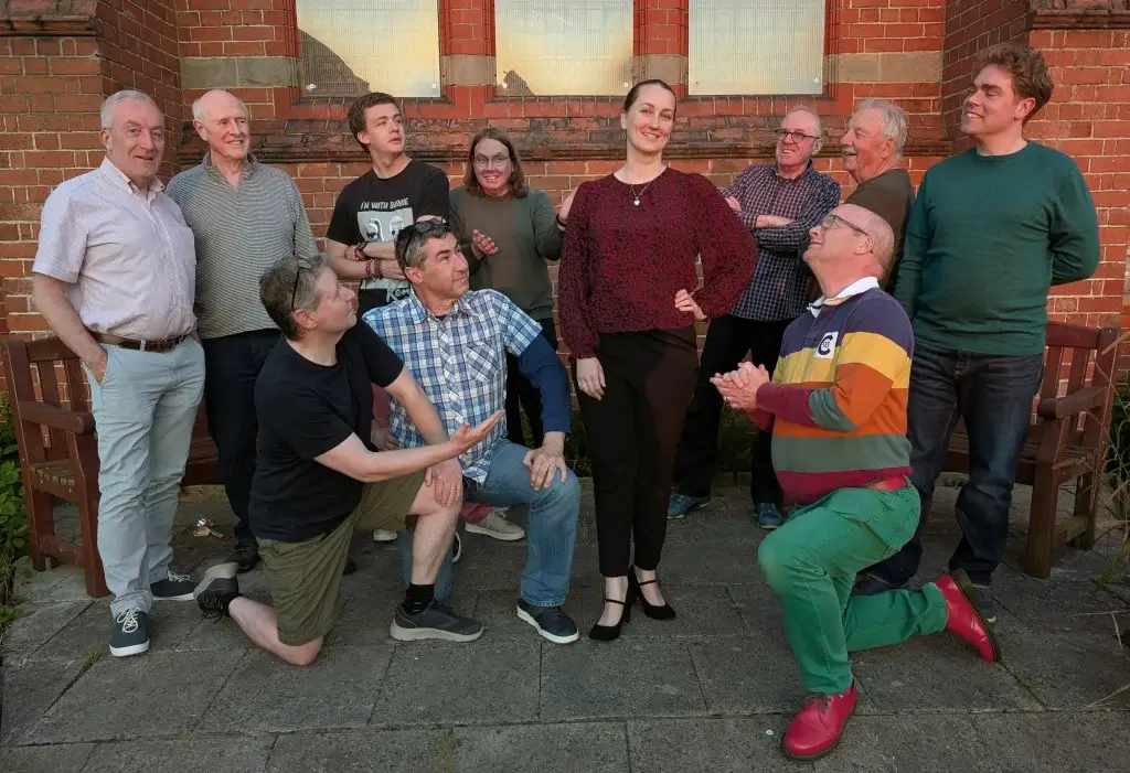 A group of people posing playfully outdoors, with some kneeling and mimicking actions, against a backdrop of a brick wall.