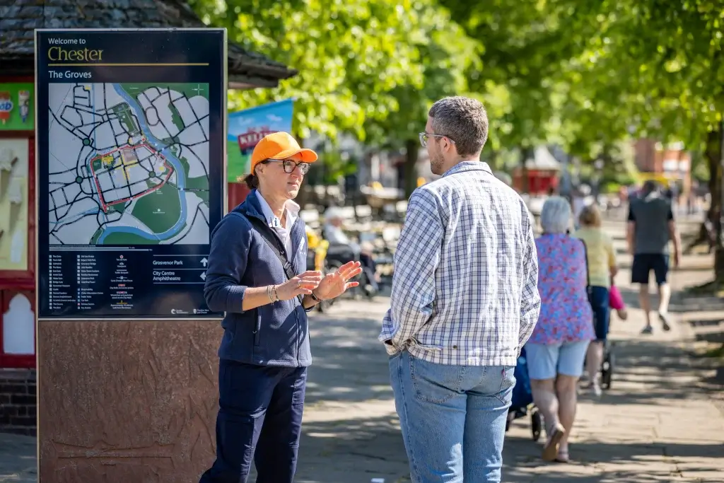 A tour guide in a blue jacket and orange cap gestures while explaining a map of Chester to a man in a checkered shirt.