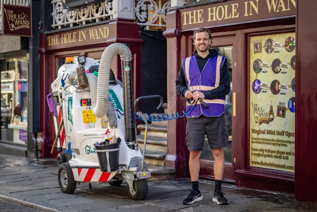A worker in a purple and black vest stands beside a cleaning machine outside "The Hole in Wand," a mini golf venue.