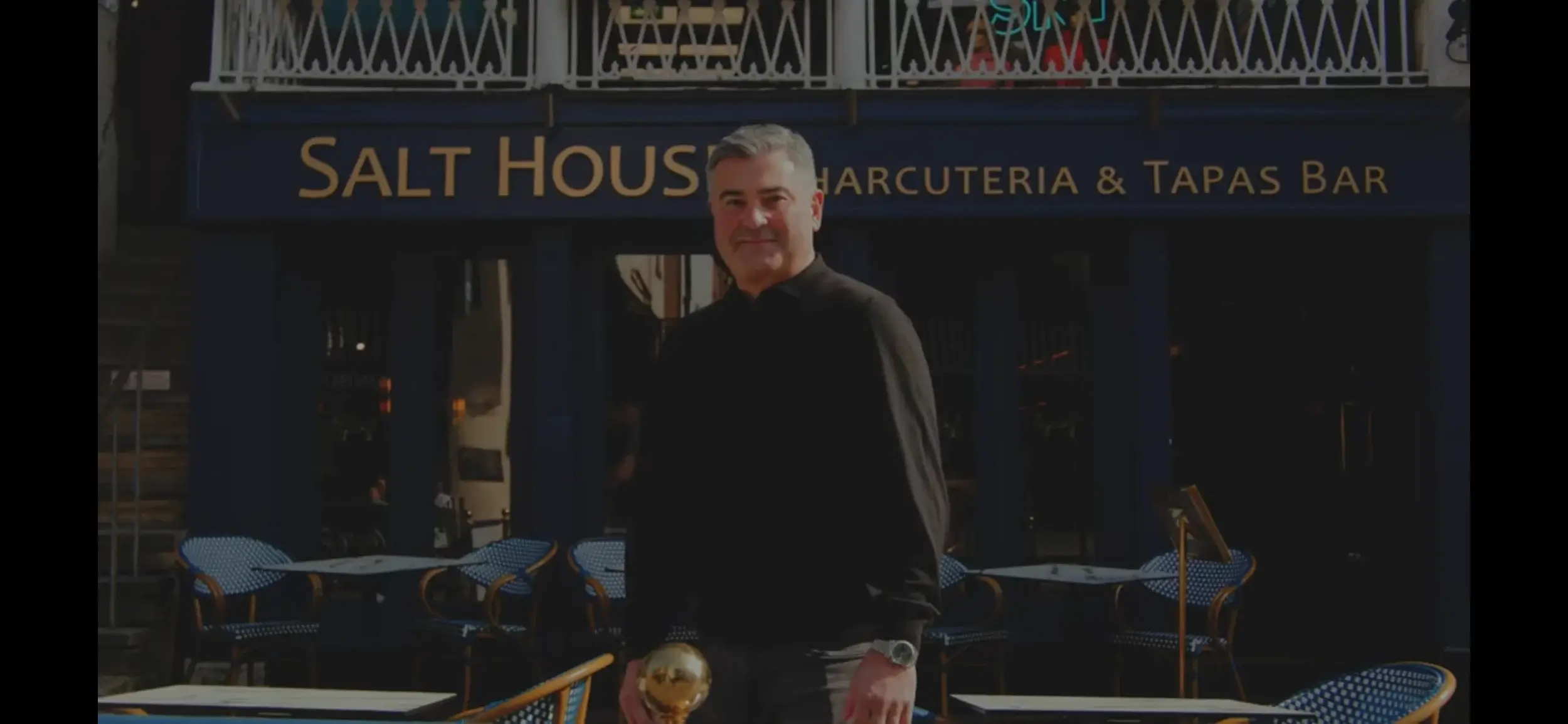 A person stands outside "Salt House," a charcuterie and tapas bar, holding a golden object, with outdoor seating in the background.