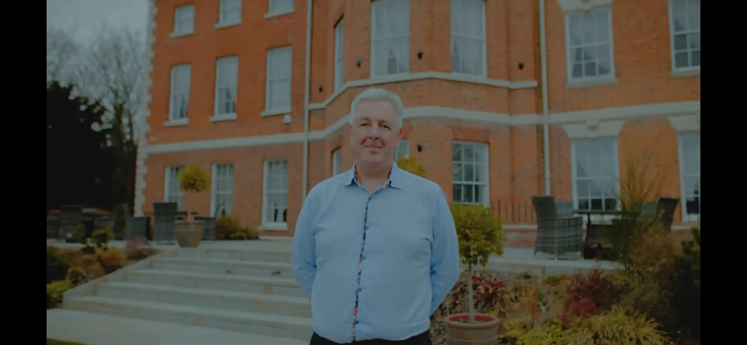 A man stands outdoors in front of a large, elegant brick building, surrounded by greenery and patio furniture.