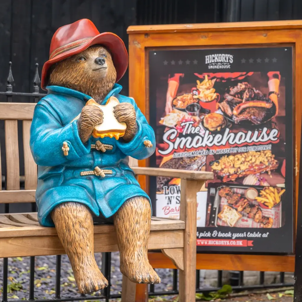 A statue of Paddington Bear, wearing a blue coat and red hat, sits on a bench eating a sandwich beside a Smokehouse restaurant sign.