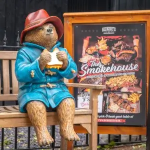 A statue of Paddington Bear, wearing a blue coat and red hat, sits on a bench eating a sandwich beside a Smokehouse restaurant sign.
