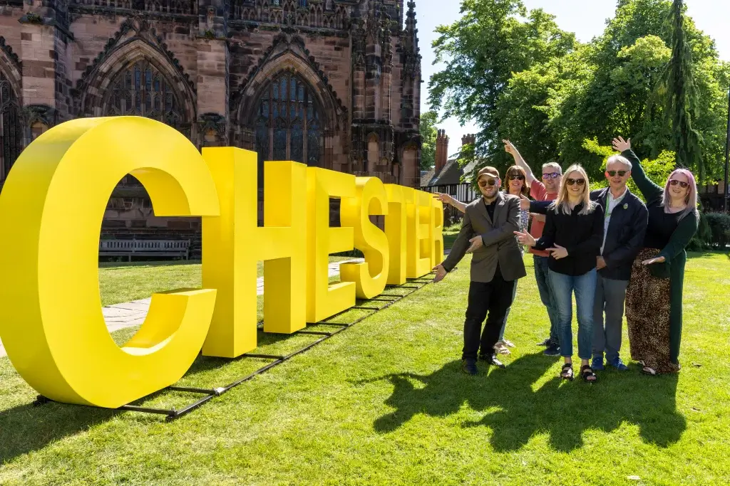 large yellow letter spelling out 'Chester' on grass outside Chester Cathedral with a group of people standing to the right