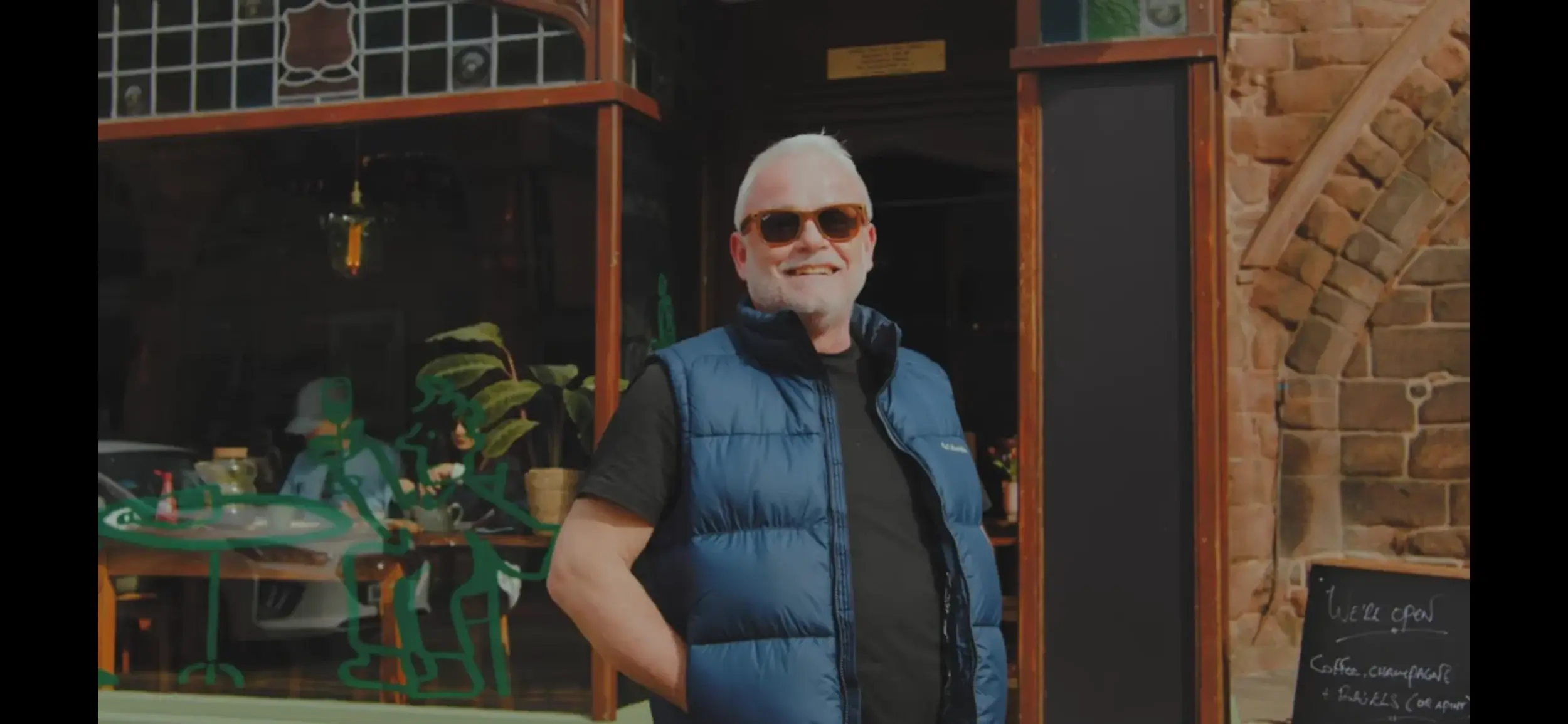 A man in a blue vest stands outside a cozy café with hanging plants and a blackboard sign that reads "We're open."
