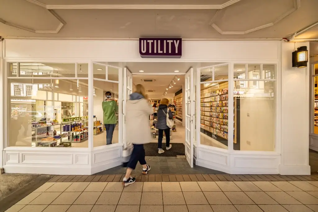 Entrance of "Utility" shop, featuring glass doors and shelves stocked with colorful products; customers entering and browsing.