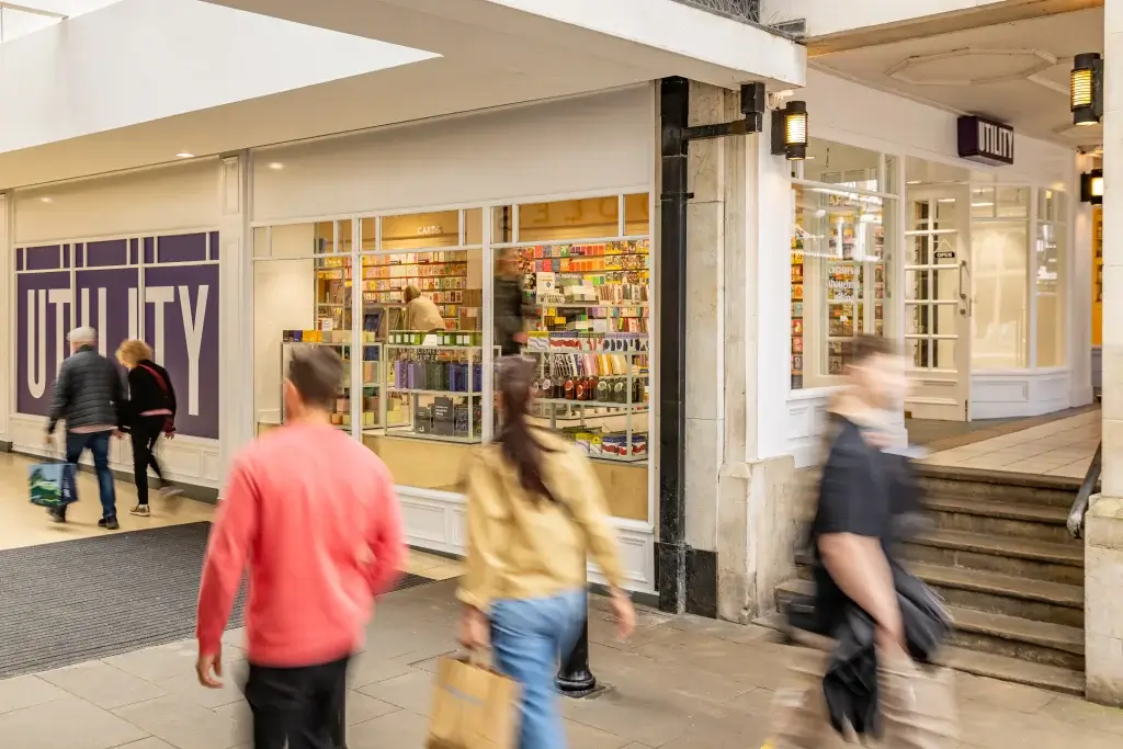 Shoppers walk past a store named "Utility," showcasing colorful cards and gifts in its window display. A busy shopping arcade scene.