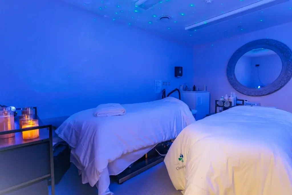 Two massage beds in a dim blue-lit spa room with lit candles, folded towels, product bottles on a trolley and a large round wall mirror.