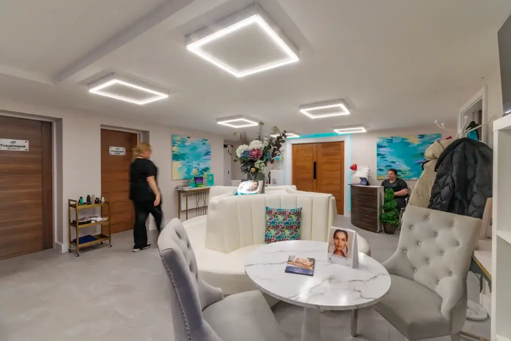 Spa reception with central curved white sofa, marble tabletop and tufted grey chairs under square LED ceiling lights.