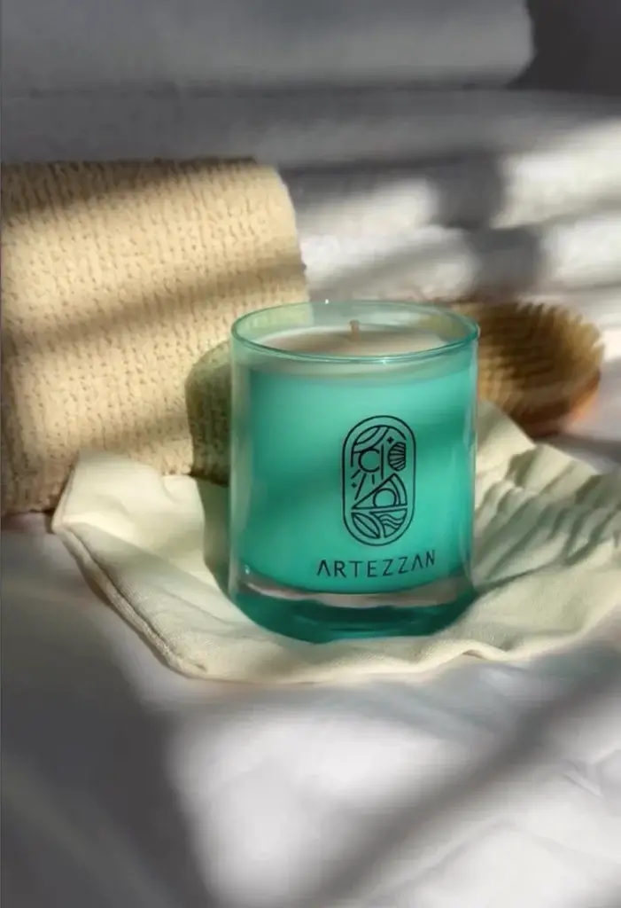 A teal candle with an 'Artezzan' logo sits on a cream cloth, surrounded by soft towels and a texture brush, illuminated by sunlight.