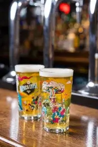 Two colorful beer glasses on a wooden bar, featuring vibrant designs, with a blurred background of bar equipment and bottles.