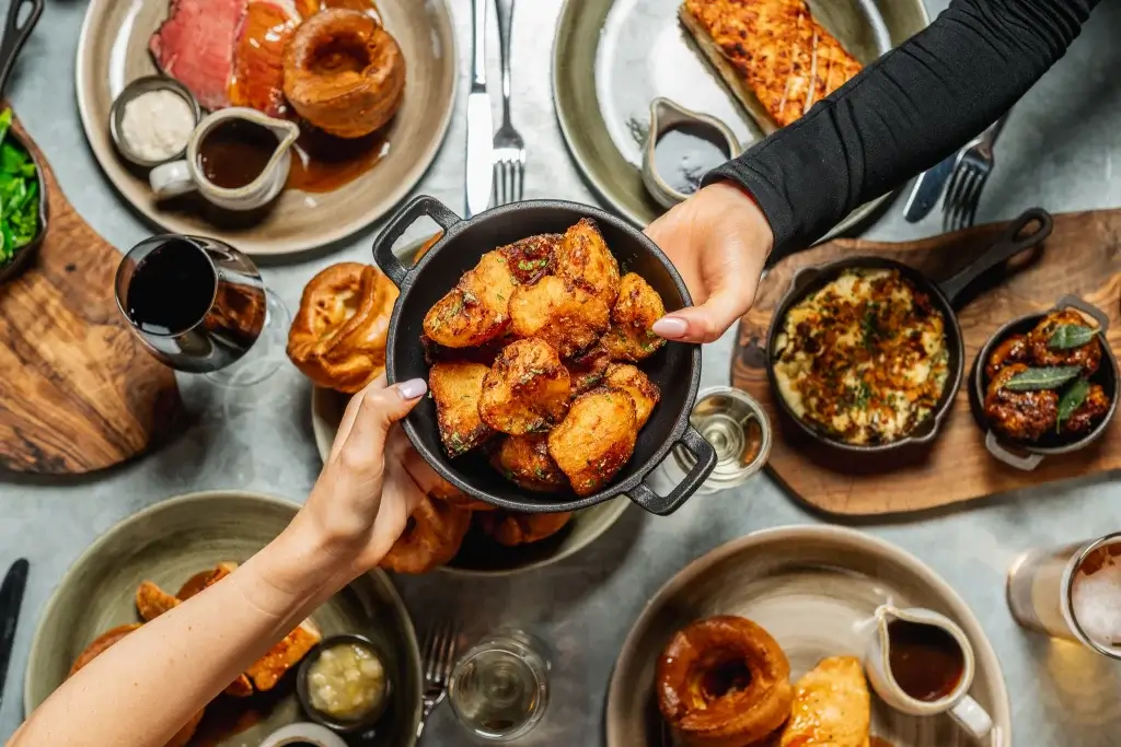 A top-down view of a feast featuring roasted meats, Yorkshire puddings, vegetables, and drinks, with hands serving and sharing food.