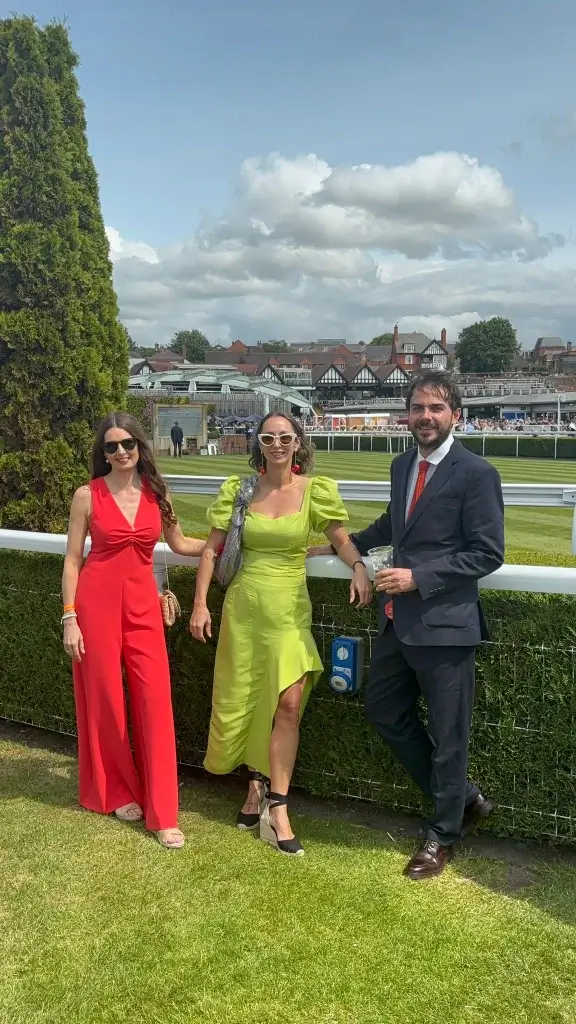 Three elegantly dressed individuals pose together on a sunny day at an event, with a landscaped background and spectators in view.