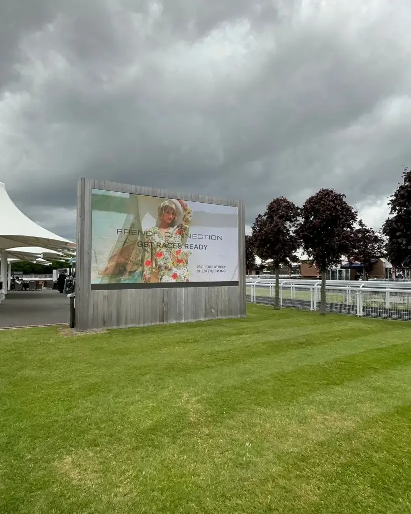 A large outdoor advertisement for "French Connection" with colorful floral designs, set against a cloudy sky and green grass.