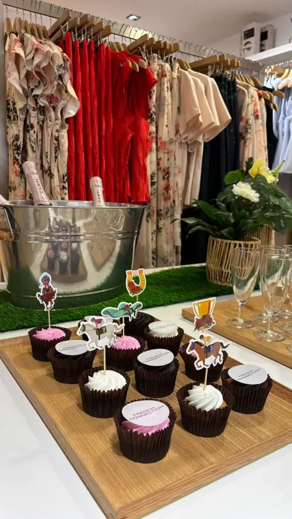 A display of cupcakes with whimsical horse-themed toppers, alongside a chilled champagne bucket and colorful dresses hanging in a boutique.