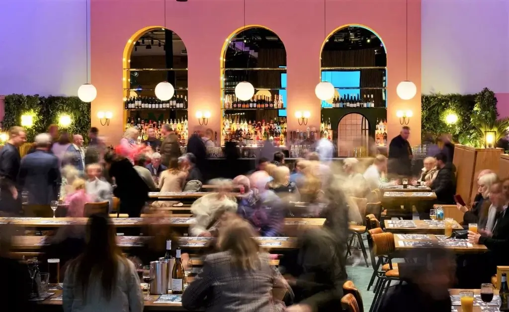A bustling restaurant scene filled with patrons, wooden tables, and a vibrant bar adorned with bottles and stylish lighting.