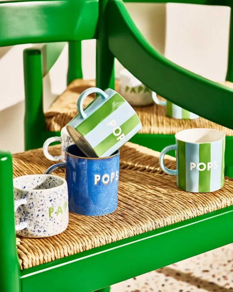 A colorful arrangement of ceramic mugs on a green chair, featuring varied designs and the word "POPS" prominently displayed.