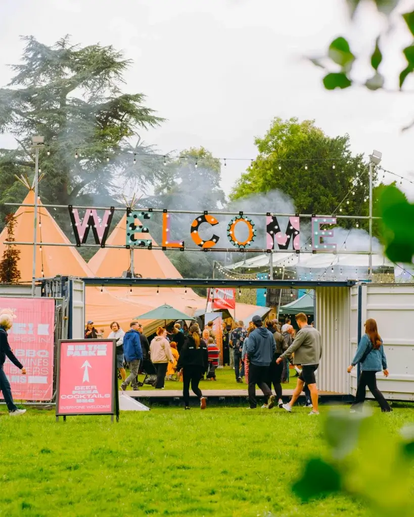 A vibrant outdoor festival entrance with a decorative "WELCOME" sign, crowds, food stalls, and colorful tents in a lush green park.