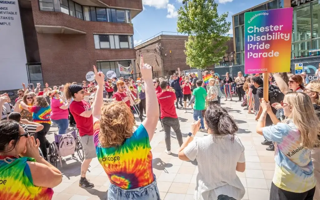 A vibrant crowd celebrates at the Chester Disability Pride Parade, dancing under sunny skies and holding colorful signs.