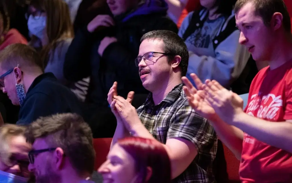 A crowd of people in a dimly lit venue, some clapping, seated in rows, showcasing various expressions and attire.