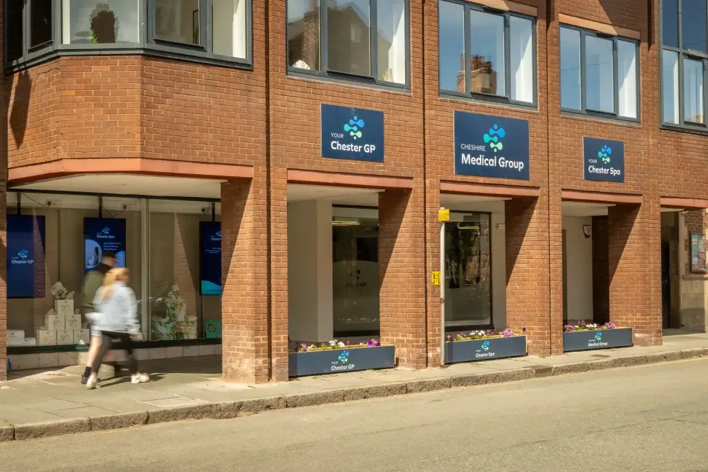 Exterior of a brick building featuring signs for Chester GP, Cheshire Medical Group, and Chester Spa, with pedestrians walking by.