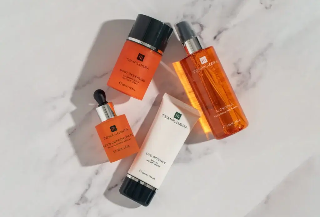 Four Templespa skincare products on a marble surface: serums, a moisturiser, and a toner in bright orange and white packaging.