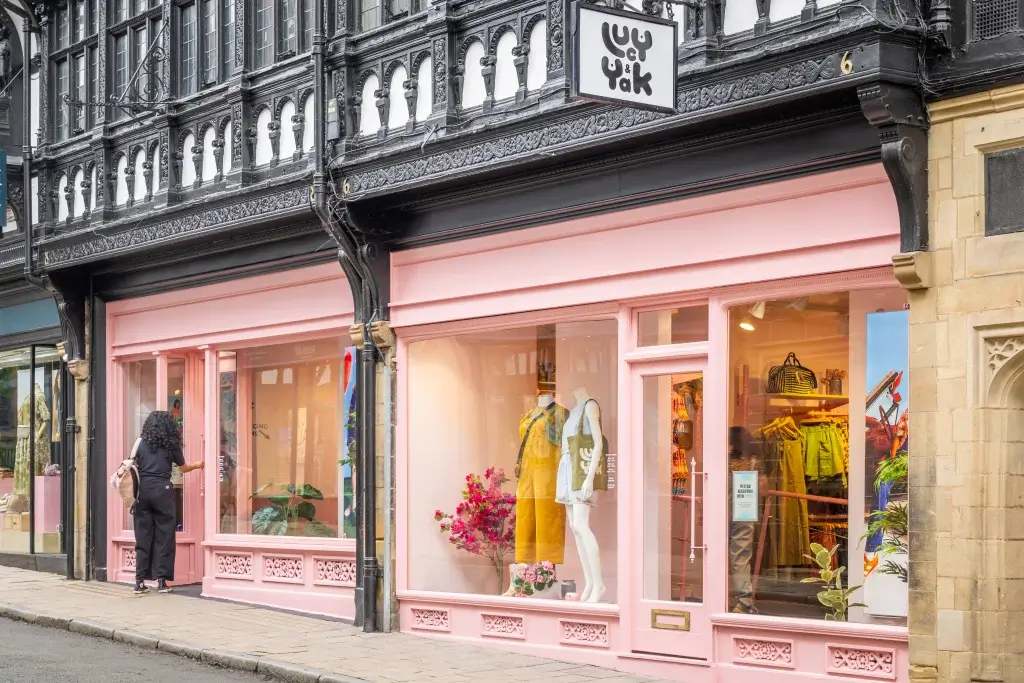 A woman with curly hair approaches a pink storefront displaying vibrant clothing and floral accents, featuring the "Lucy & Yak" logo above.