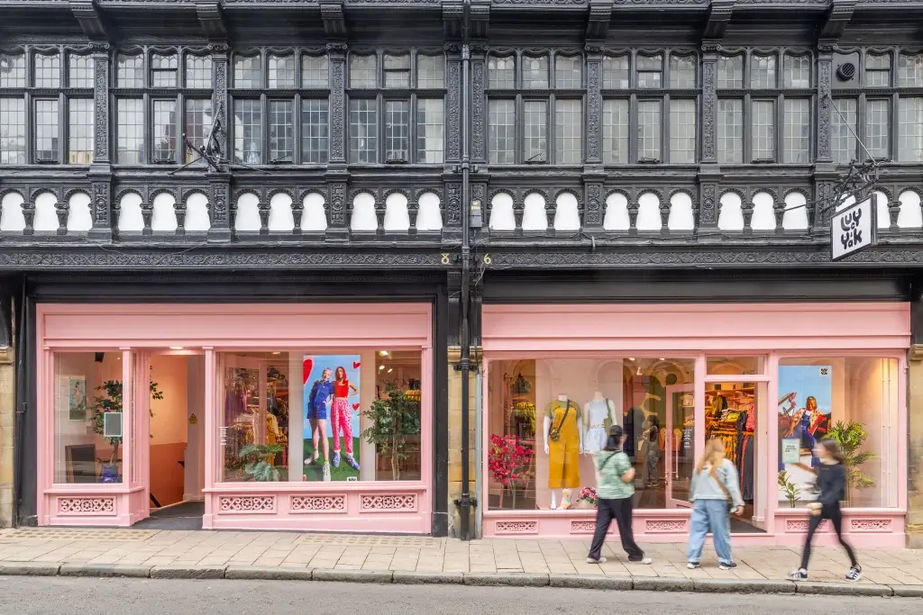 Street view of a pink storefront showcasing clothing and mannequins, framed by an ornate black and white building, with pedestrians walking by.