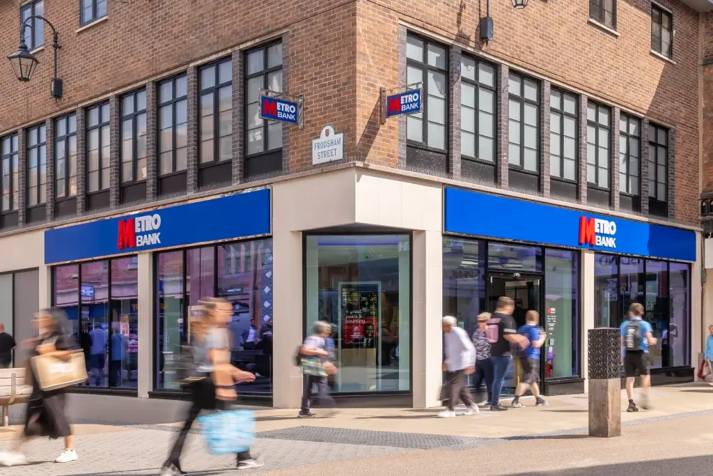 A busy street scene featuring the Metro Bank storefront, with pedestrians walking past on a sunny day.