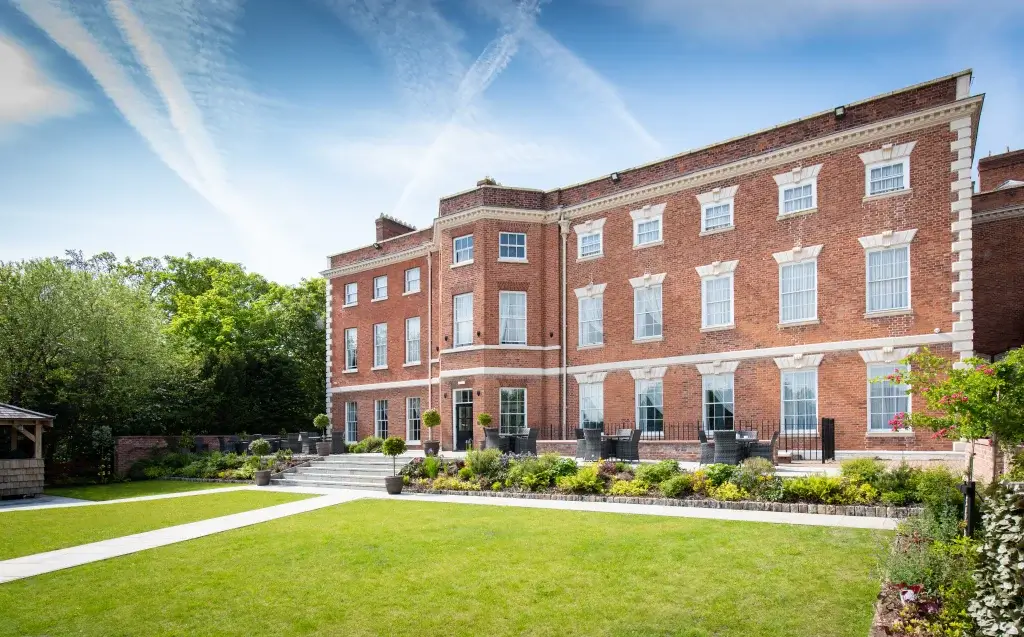 Elegant red brick building with white window accents, surrounded by a landscaped garden and blue sky.