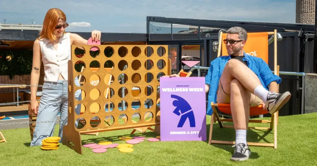 one person is playing a wooden game whilst the other sits in an chair that says Aperol. both perched on artificial grass. a sign that says wellness week can also be seen