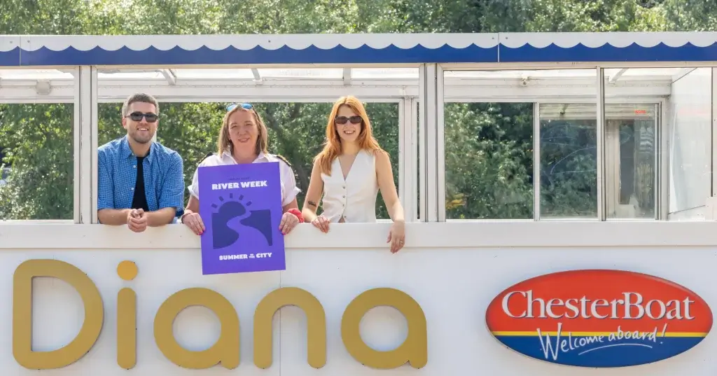 three people wearing sunglasses look out from a boat towards camera holding a sign that says river week. the words Chester Boat and Diana can be seen on the boat