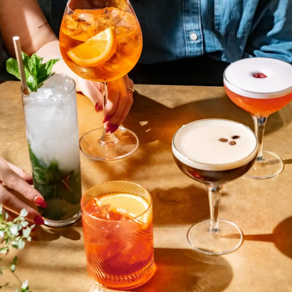 A selection of colorful cocktails on a table, including a mojito, espresso martini, and spritz, with hands reaching for drinks.
