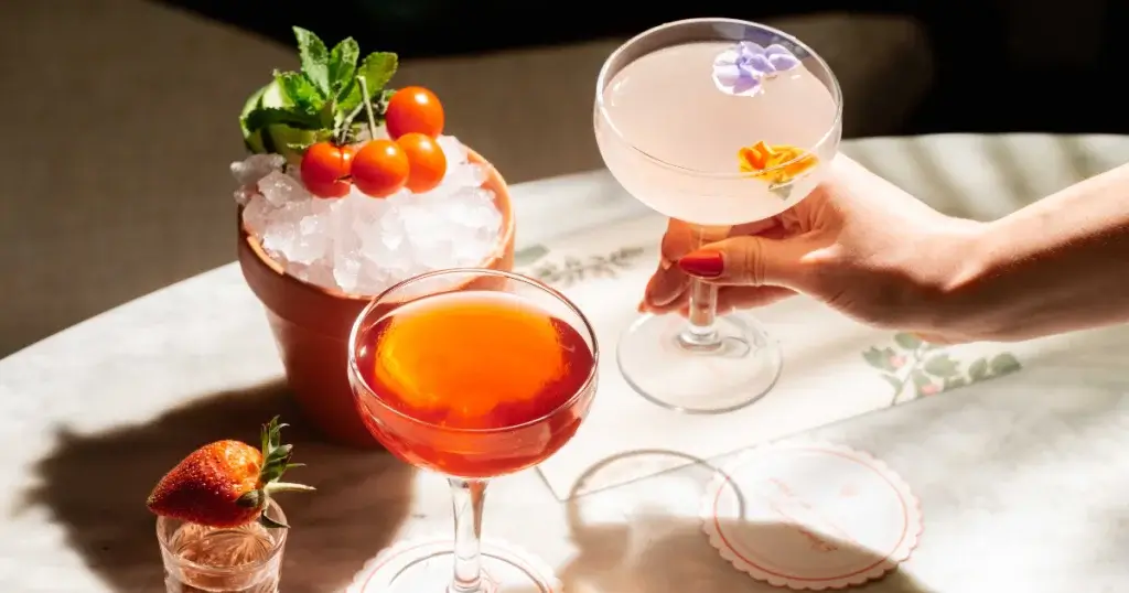 A hand holds a cocktail with floral garnish beside a vibrant red drink, fresh tomatoes, ice, and a strawberry on a marble table.