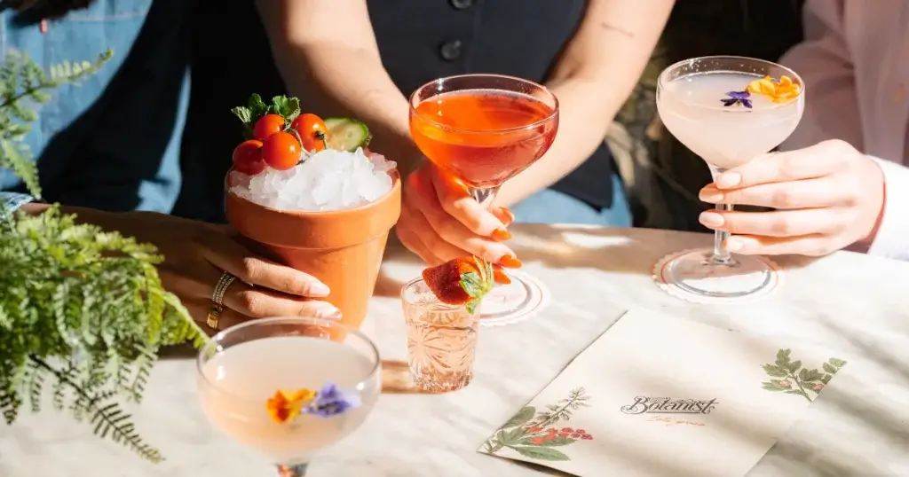 A vibrant display of cocktails on a table, including a terracotta pot drink, adorned with herbs and flowers, with two hands holding drinks.