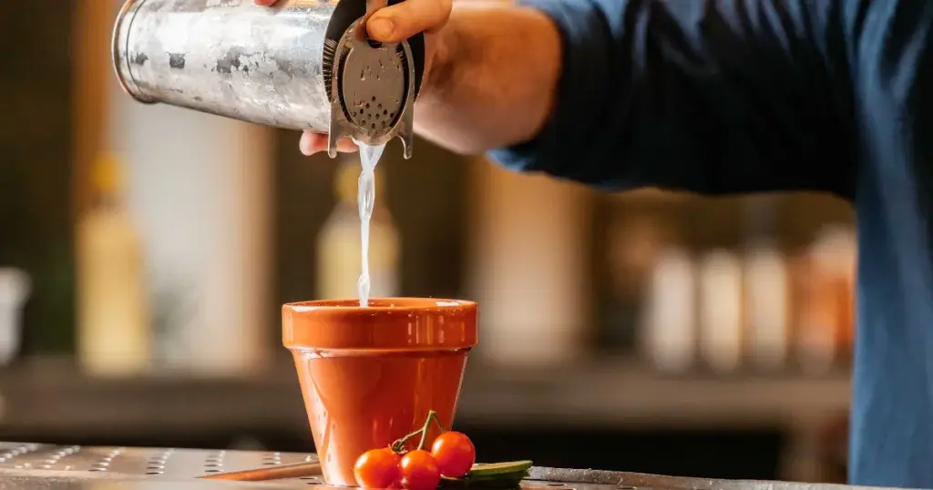 A bartender pours a clear liquid from a cocktail shaker into a small terracotta pot, surrounded by cherry tomatoes and herbs.