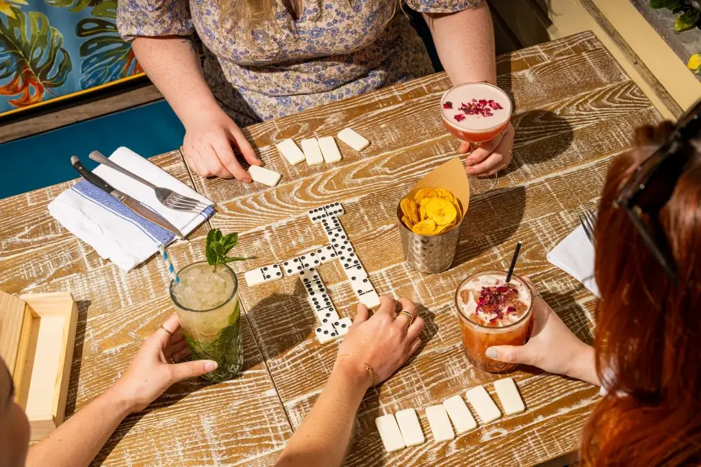 A tabletop scene of friends playing dominoes with drinks and snacks, surrounded by a vibrant, tropical backdrop.