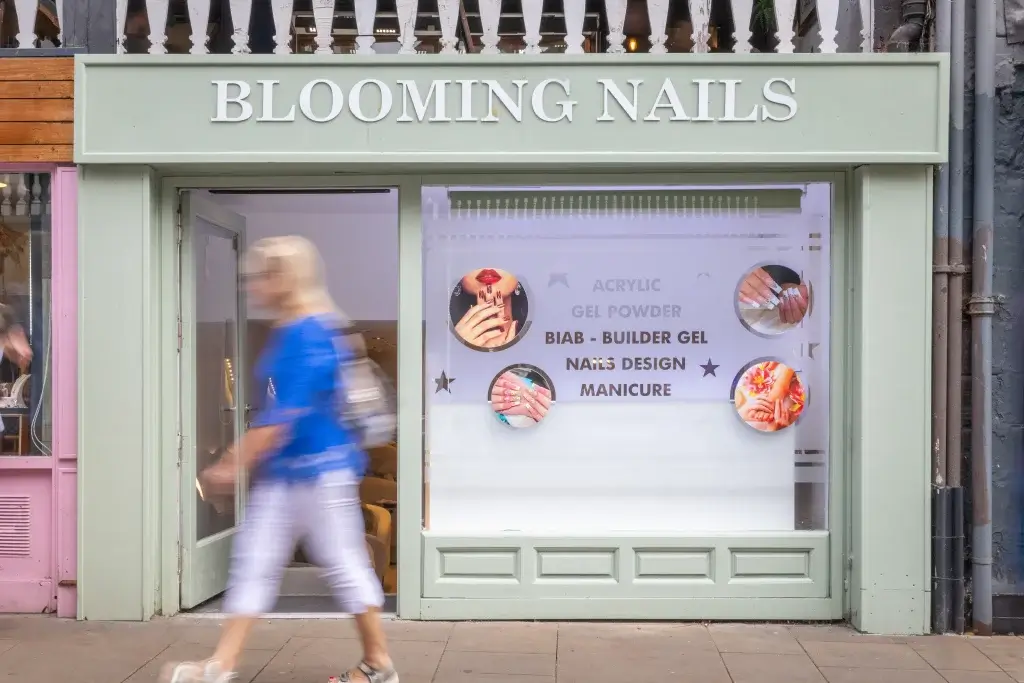 A storefront sign reading "Blooming Nails" displays nail services with images of manicured hands as a woman walks by.
