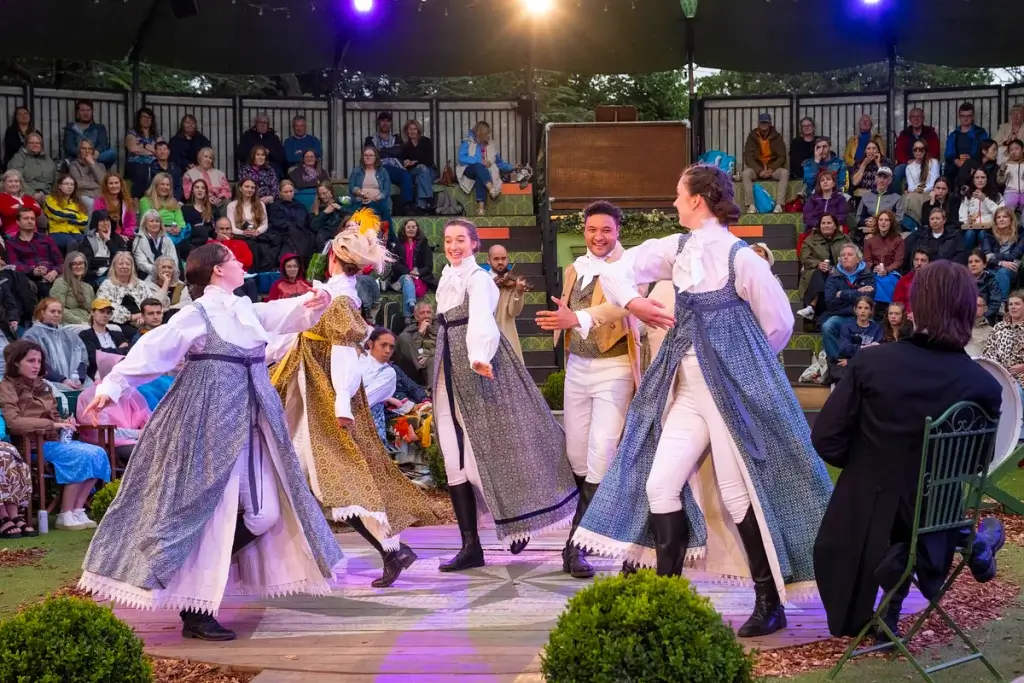 A lively dance performance featuring costumed dancers in colorful attire, surrounded by an engaged audience in an outdoor theater setting.