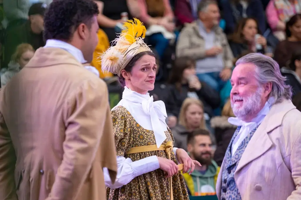 Three individuals in historical costumes converse, with an audience of spectators in the background. One wears a feathered hat and a floral dress.