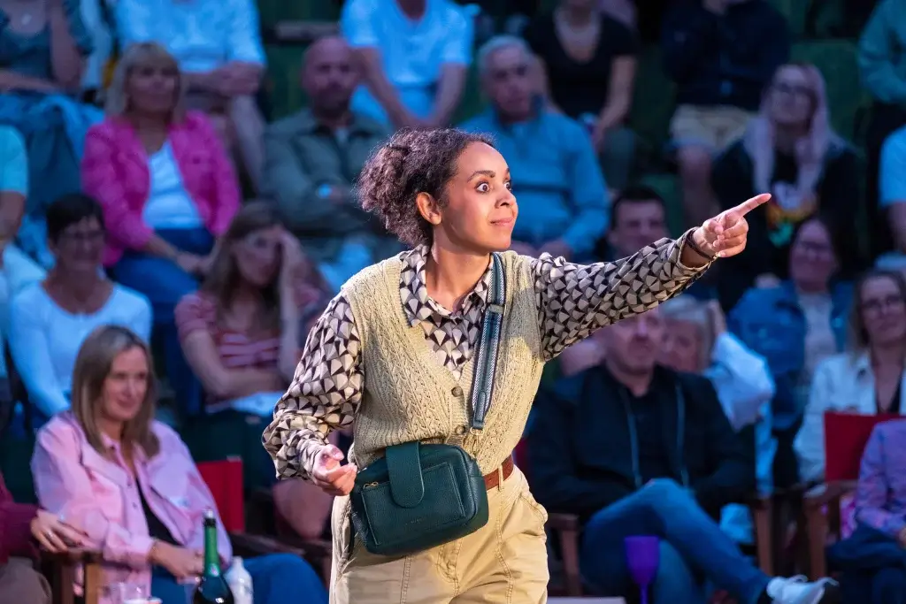 A performer passionately gestures while wearing a patterned shirt, knitted vest, and holding a green handbag, surrounded by an engaged audience.