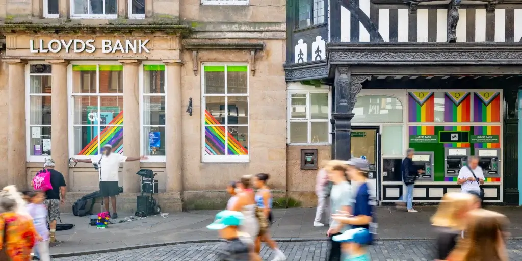 Lloyds Bank with rainbow-striped windows and ATMs under a timbered façade, busy street with blurred pedestrians.