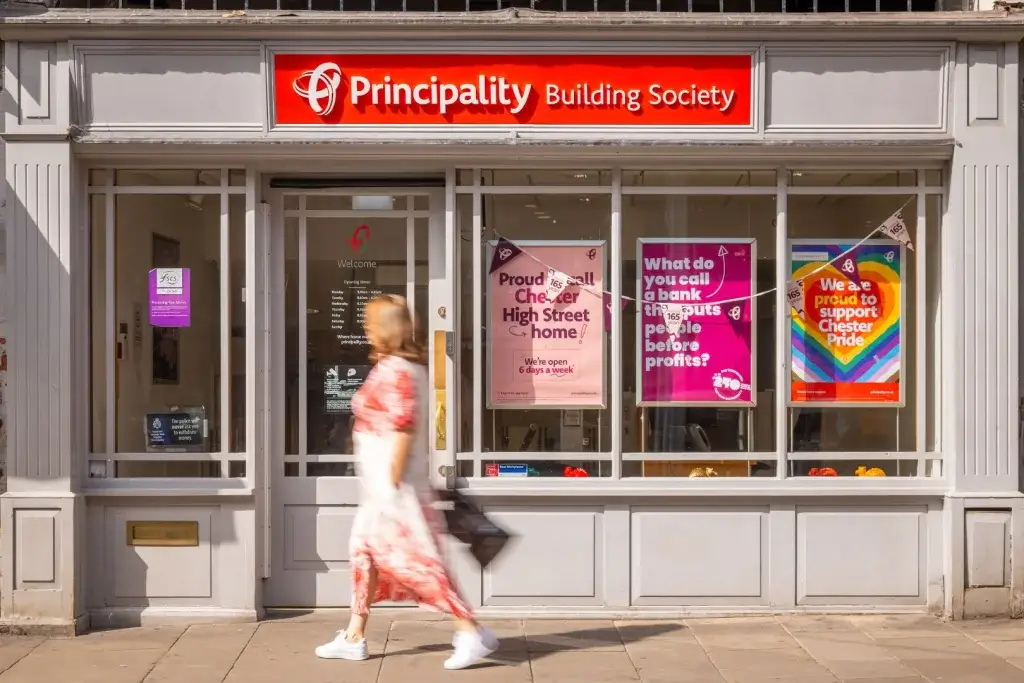 Principality Building Society branch with red sign, colourful pride and promo posters in windows and a blurred woman walking past.