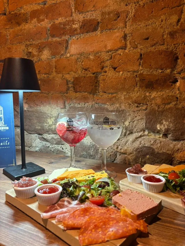 Charcuterie board with meats, pâté, cheeses and dips beside two gin cocktails on a wooden table against a brick wall.
