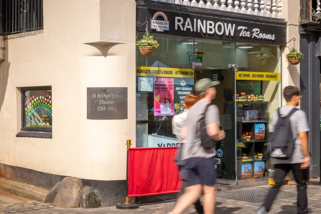 Corner Rainbow Tea Rooms storefront with hanging baskets, rainbow window display and blurred pedestrians.