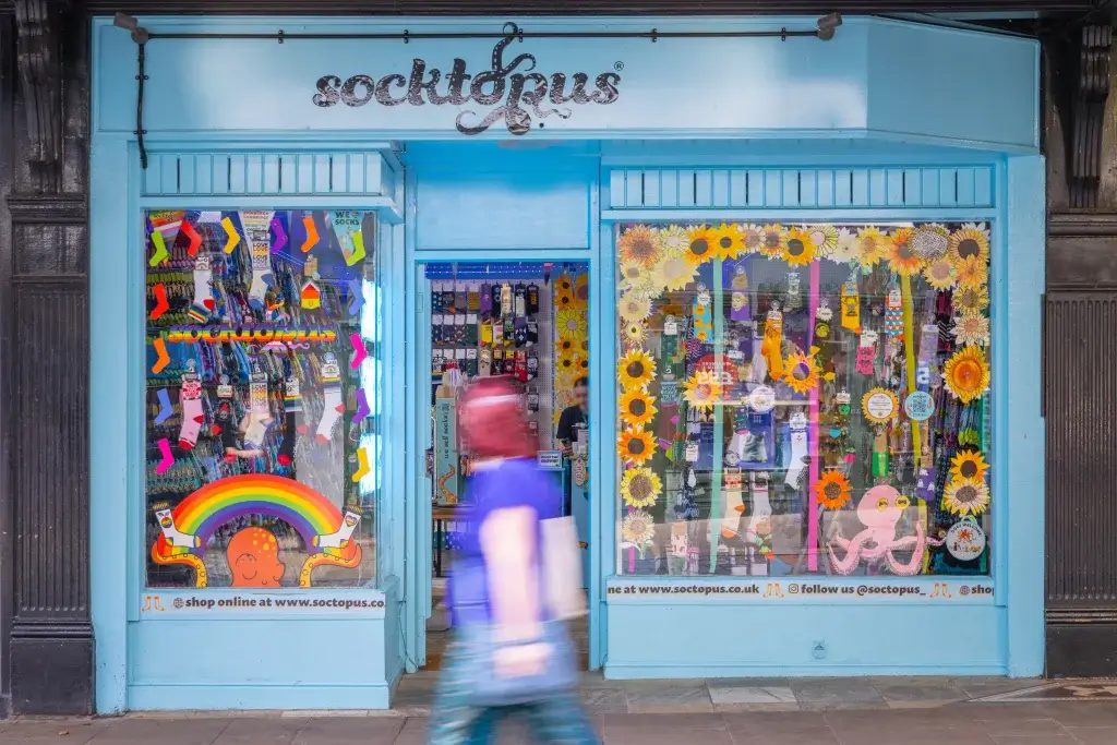Turquoise socktopus shopfront with colourful sock displays, rainbow and sunflower window art and a blurred pedestrian.